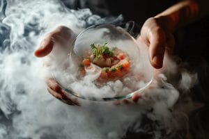 fotografía de manos sosteniendo un plato bajo una campana de cristal llena de humo