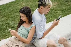 Dos jóvenes sentados de espaldas mirando sus teléfonos en un parque