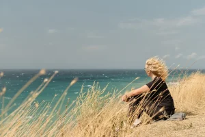 mujer sentada de espaldas en una colina frente al mar, contemplando el horizonte en un entorno de naturaleza tranquila.